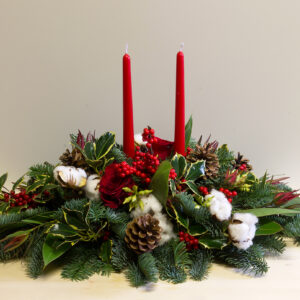 Elegante centrotavola natalizio con due candele rosse, rami di abete, bacche, pigne e cotone, ideale per decorazioni e regali di Natale.