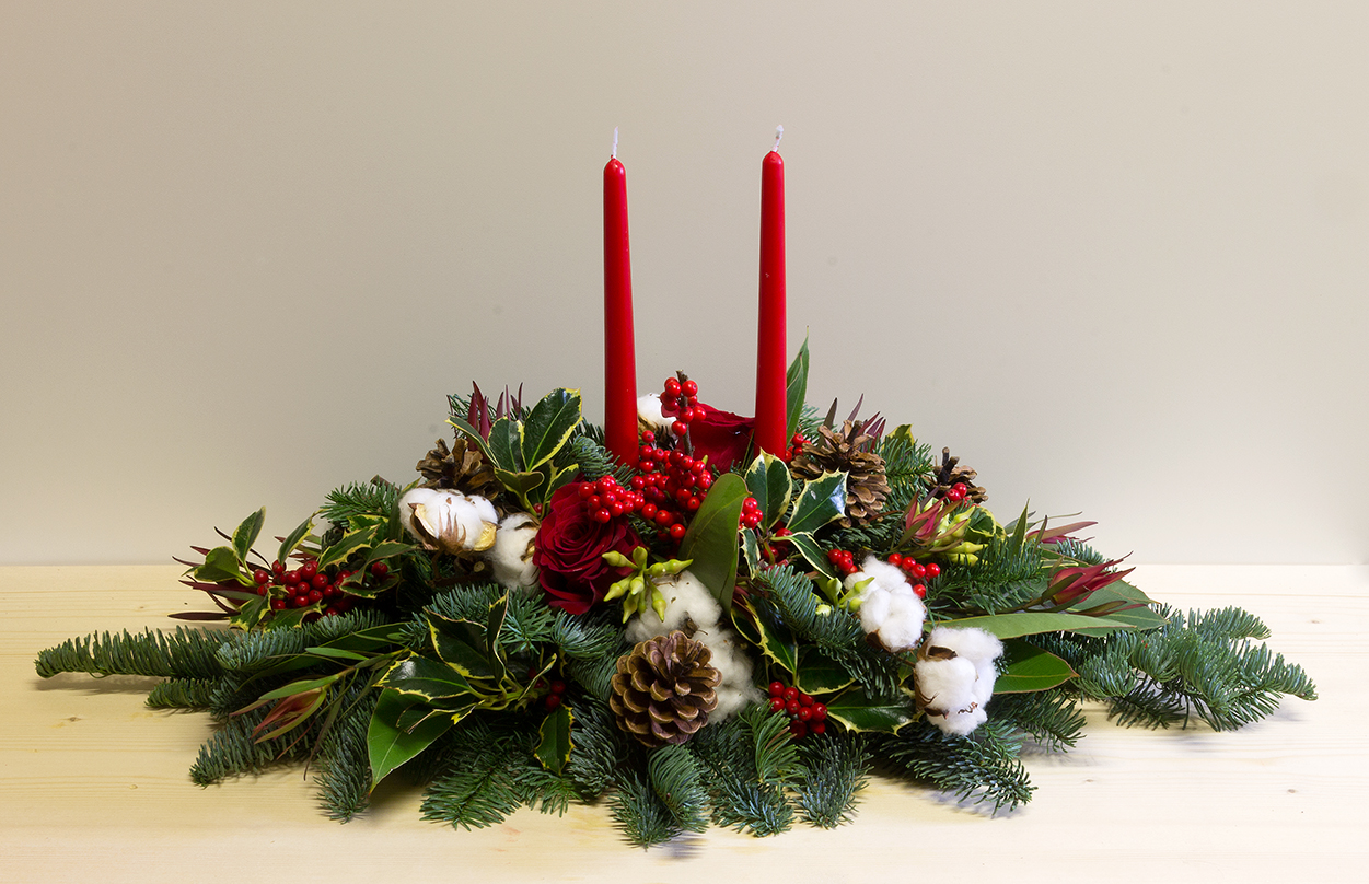 Elegante centrotavola natalizio con due candele rosse, rami di abete, bacche, pigne e cotone, ideale per decorazioni e regali di Natale.