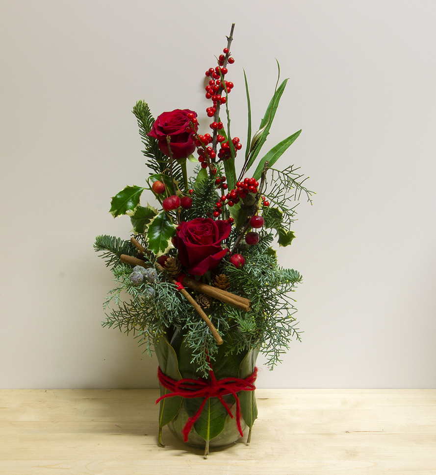 Composizione floreale verticale "Merry Christmas" con rose rosse, abete, bacche, pigne e cannella, legata con nastro rosso.