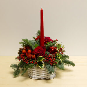 Centrotavola natalizio Happy Christmas con candela rossa, rose rosse, bacche, abete e pigne in cestino tondo bianco.