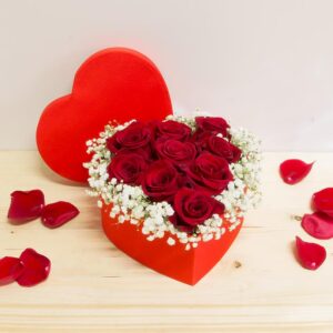 Scatola a forma di cuore di colore rosso vellutato piena di rose rosse fresche e fiori di nebbiolina bianca su un tavolo di legno chiaro con petali sparsi.