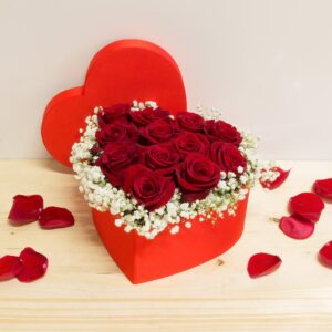 Scatola rossa a forma di cuore con 12 rose rosse fresche e nebbiolina bianca su piano in legno.