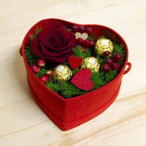 Box regalo vellutata a forma di cuore rosso contenente una rosa bordeaux, tre cioccolatini Ferrero Rocher dorati, cuori decorativi e rametti verdi su un tavolo in legno chiaro.