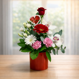 Composizione floreale Love me tender in vaso rosso con rosa rossa, orchidee bianche, gerbere rosse e bianche, garofani rosa e un cuore decorativo traforato.