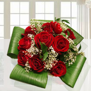 Bouquet di 9 rose rosse fresche con foglie verdi di felce in un vaso di vetro cilindrico su fondo luminoso.