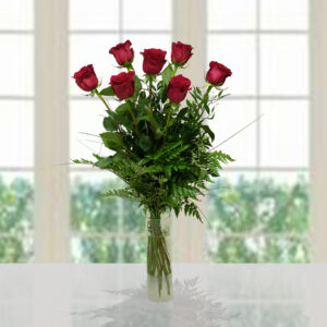 Bouquet romantico di 7 rose rosse a stelo lungo con abbondante verde decorativo in un vaso di vetro cilindrico trasparente.