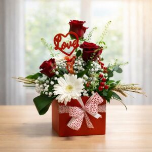Flower box quadrata rossa Batticuore con tre rose rosse, una gerbera bianca, gipsofila, bacche rosse, spighe e un topper rosso con la scritta Love.