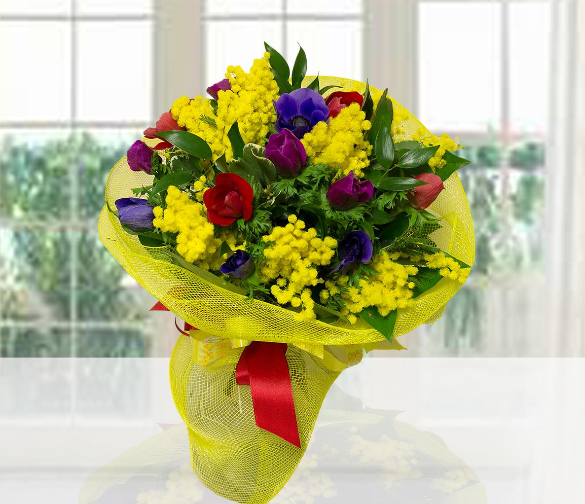 Ricco bouquet di anemoni colorati (blu, viola, rossi) e mimosa gialla, confezionato con rete gialla e fiocco rosso.
