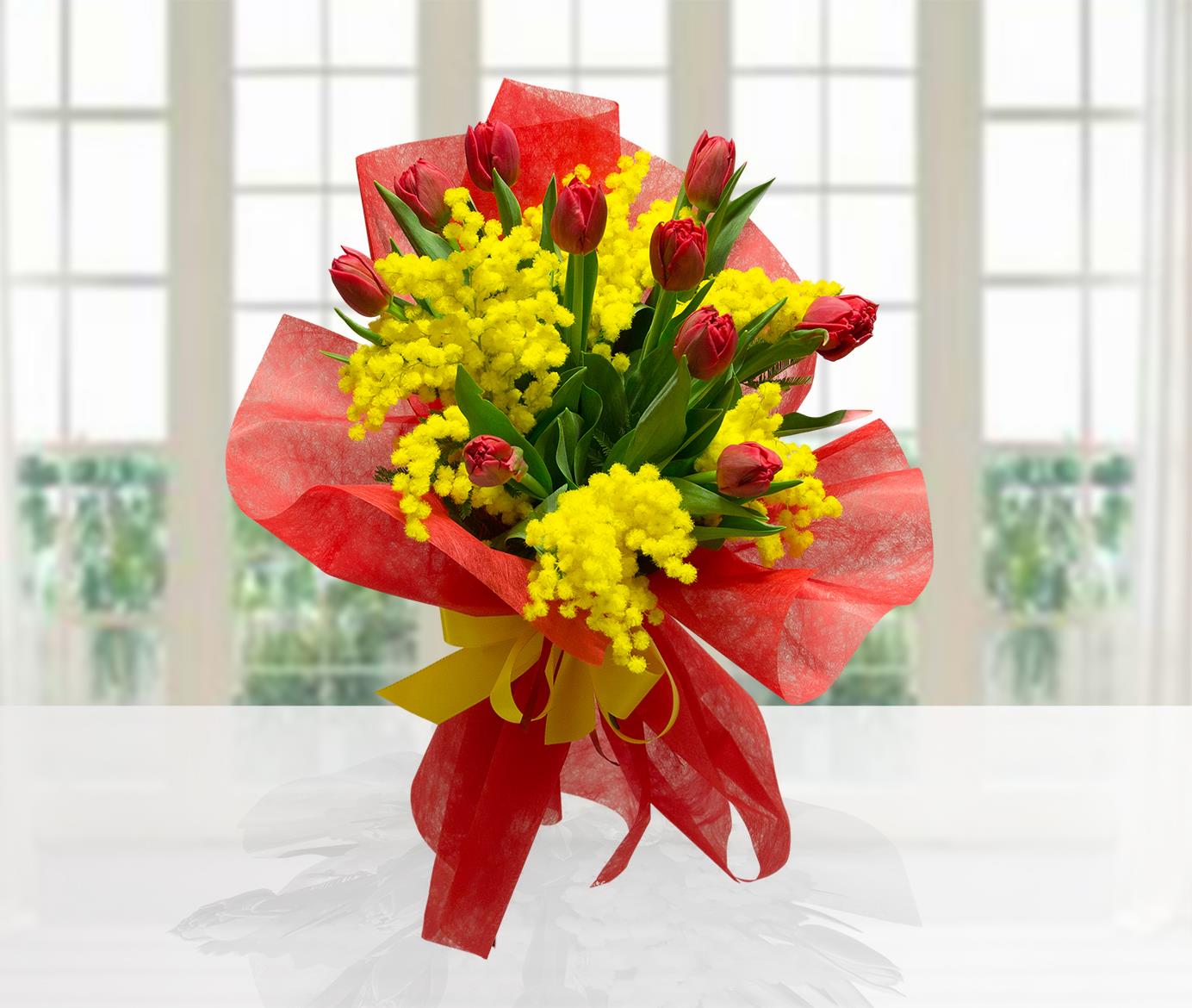 Mazzo di tulipani rossi e mimosa gialla, confezionato con carta velina rossa e fiocco giallo su sfondo luminoso.