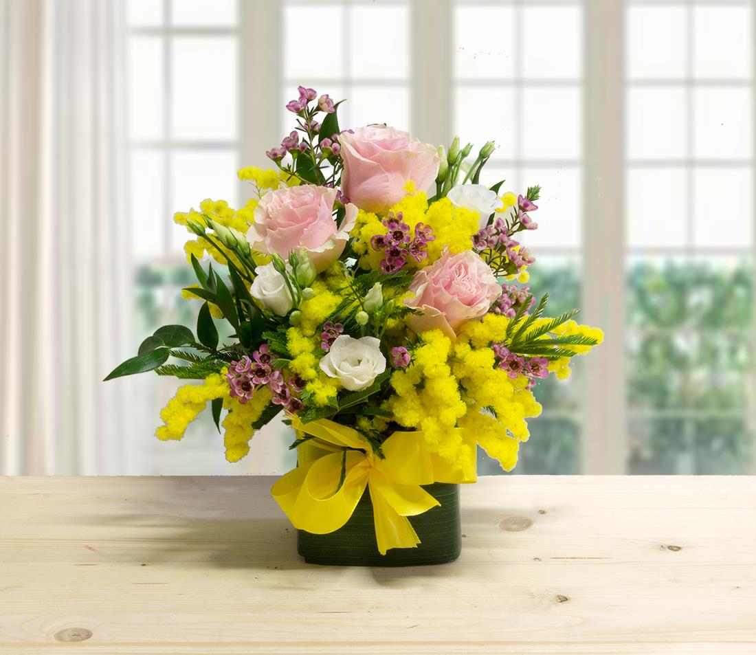 Composizione floreale Rose Little Woman con rose rosa, mimosa gialla e lisianthus bianchi in vaso quadrato con fiocco giallo.